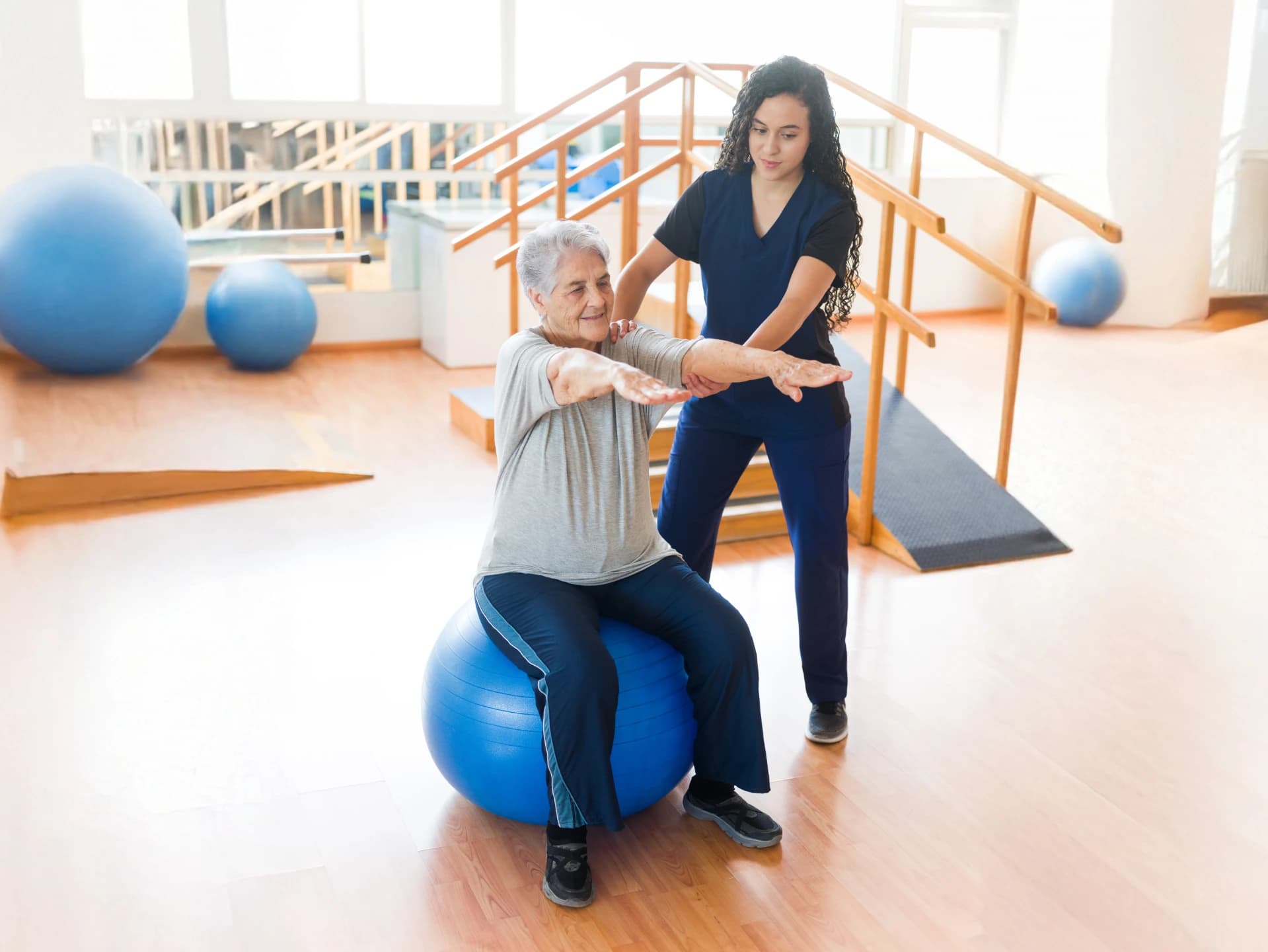 Seniorin dehnt ihre Arme auf einem Gymnastikball neben einer Ergotherapeutin